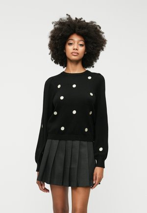 Black sweater with metallic gold polka dots, round neckline, long sleeves. Paired with a pleated dark skirt for a complete look.