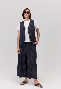 Granatowa kamizelka w prążki noszona na białej koszulce, w zestawieniu z szerokimi spodniami i czarnymi sandałami. Kamizelka ma guziki i kieszenie.