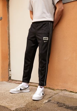 Personne portant un pantalon de jogging noir Nike avec des bandes blanches sur les côtés et des baskets blanches Nike, debout contre un mur orange et un sol en béton.