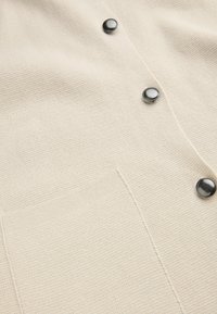 Stickad cardigan i krämfärg med en texturerad yta, med tre runda svarta knappar och en framficka.