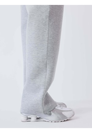 Jambes en pantalon de survêtement gris clair couvrant la majeure partie de chaussures de sport blanches et grises avec des semelles à coussin d'air visibles, debout sur un sol blanc.