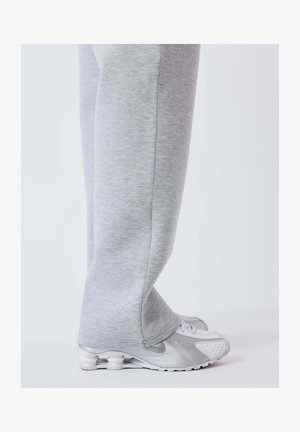 Jambes en pantalon de survêtement gris clair couvrant la majeure partie de chaussures de sport blanches et grises avec des semelles à coussin d'air visibles, debout sur un sol blanc.
