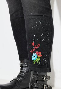 Pantalones vaqueros de mezclilla negra con manchas de pintura blanca, que presentan estampados florales coloridos y el texto "XOXO" en la parte inferior de la pierna. Botas negras con hebilla y tachuelas.