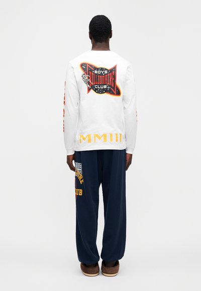 Άσπρο μακρυμάνικο πουκάμισο με πολύχρωμο γραφικό σχέδιο στην πλάτη· σκούρο μπλε παντελόνι με κείμενο σε διάφορα χρώματα· μαλακό ύφασμα και άνετη γραμμή.