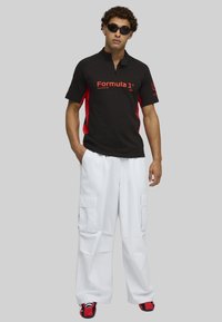 Musta polo, jossa on punaisia yksityiskohtia, vetoketjutakki ja "Formula 1" -logo, yhdistettynä löysiin valkoisiin cargo-housuihin ja punaisiin lenkkareihin.
