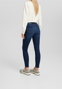 Modré skinny džíny s vysokým pasem, hladkou texturou a pěti kapsami, doplněné krémovým zkráceným svetrem a šedými teniskami.