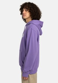 Pullover hoodie in lavender, made of soft fabric. Features a front pocket, adjustable drawstring hood, and logo accents on the chest.