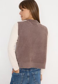 Frau trägt eine ärmellose, fuzzy mauve Weste über einem langärmligen cremefarbenen Oberteil und blauen Jeans, mit den Händen in den hinteren Hosentaschen, von hinten blickend.