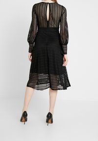 Černé pruhované šaty s průhlednými dlouhými rukávy, výstřihem ve tvaru klíče na zádech a rozšířeným lemem. Oblečené s černými vysokými podpatky, které mají hladkou texturu a vrstvený design.