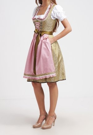 Kvinde iført en traditionel dirndl-kjole med grøn krop, hvid bluse, pink forklæde og beige højhælede sko.