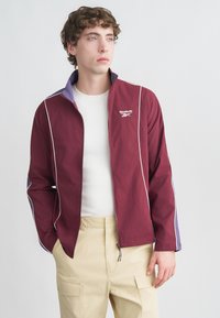 Bordowa kurtka na zamek z białymi wykończeniami, fioletowymi akcentami i logo Reebok. Lekka tkanina. Połączona z kremową koszulą i beżowymi spodniami typu cargo.