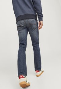Vaqueros de mezclilla azul oscuro con corte recto, costuras en contraste y dos bolsillos traseros. El calzado incluye zapatillas blancas con acentos rojos.