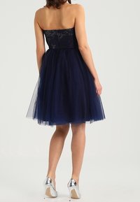 Mörkblå avskuren klänning med spetsliv och tylskjol. Parat med silvriga högklackade skor. Bakifrån visas den tighta designen.