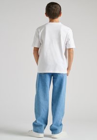 Camiseta blanca de algodón con mangas cortas, combinada con unos vaqueros azules holgados y zapatillas blancas, vista desde atrás.