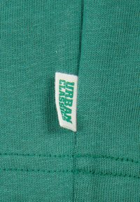 Material textil verde cu un model de tricotaj texturat, având o etichetă albă cusută pe margine cu text de marcă verde.