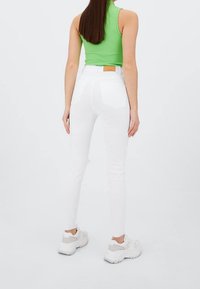Jean skinny blanc taille haute, avec poches arrière et ourlet brut. Associé à un top sans manches vert et des baskets blanches épaisses.