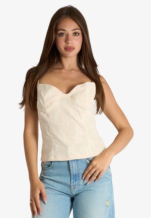 Giovane donna con lunghi capelli castani che indossa un top crema testurizzato senza spalline e jeans azzurri chiari, posando con una mano sul fianco.