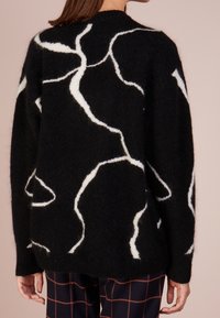 Pull noir en maille avec motif abstrait blanc, col rond et manches longues. Texture douce avec poignets et ourlet côtelés.