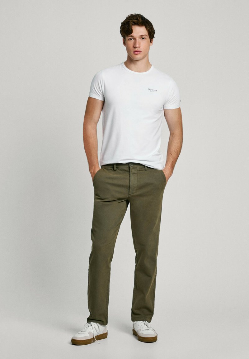 Camiseta blanca de algodón con un pequeño logo, pantalones color oliva y zapatillas blancas con suelas marrones, de pie con las manos en los bolsillos frente a un fondo liso.