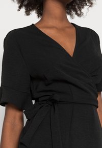 Vestido negro envolvente hecho de una tela texturizada, con mangas cortas y un detalle de cintura atada. El diseño incluye un escote en forma de V.