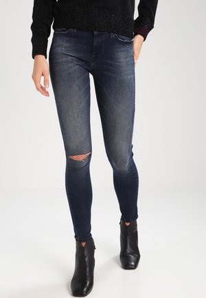 Persoon draagt donkerblauwe skinny jeans met een scheur bij de knie, gecombineerd met zwarte enkellaarzen en een zwarte trui, staand op een witte achtergrond.