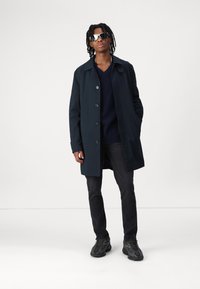 Marineblauer Trenchcoat mit Knopfverschluss, über einem dunklen Pullover getragen. Dunkle Jeans und schwarze Sneakers. Model trägt reflektierende Sonnenbrillen.