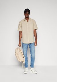 Chemise beige à manches courtes avec une poche poitrine, associée à un jean bleu clair et des baskets blanches. Les accessoires comprennent une casquette noire et un sac à dos beige.
