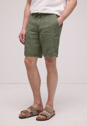 Mand iført olivengrønne linnedshorts og brune sandaler med dobbelte remme, stående med den ene hånd i lommen mod en ensfarvet baggrund.