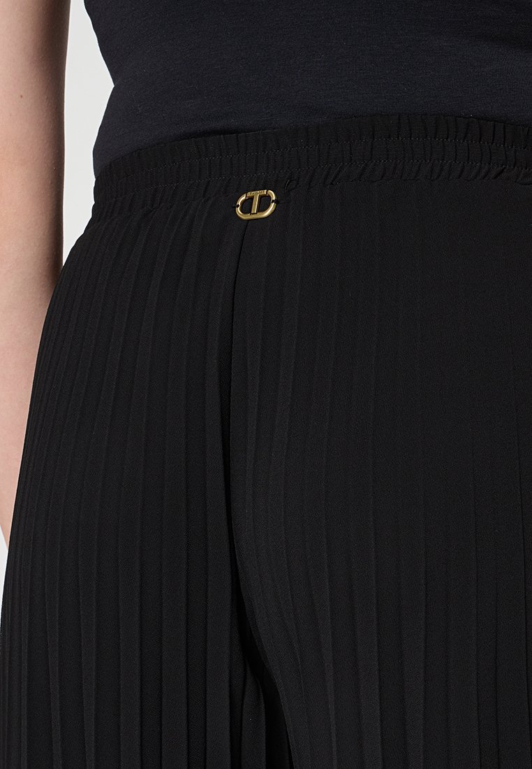 Un pantalon noir plissé avec une taille élastique et un détail en accent doré. Le tissu a un aspect lisse et texturé.