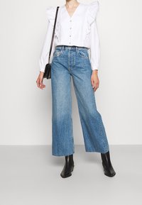 White blouse with ruffled shoulder detail and buttons, paired with blue wide-leg jeans and black ankle boots. Simple, clean design.