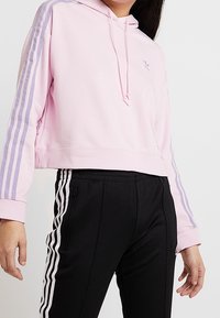 Kvinna iklädd en rosa kortärmad huvtröja med lila ränder och svarta byxor som har vita sidostripes och zip-fickor.