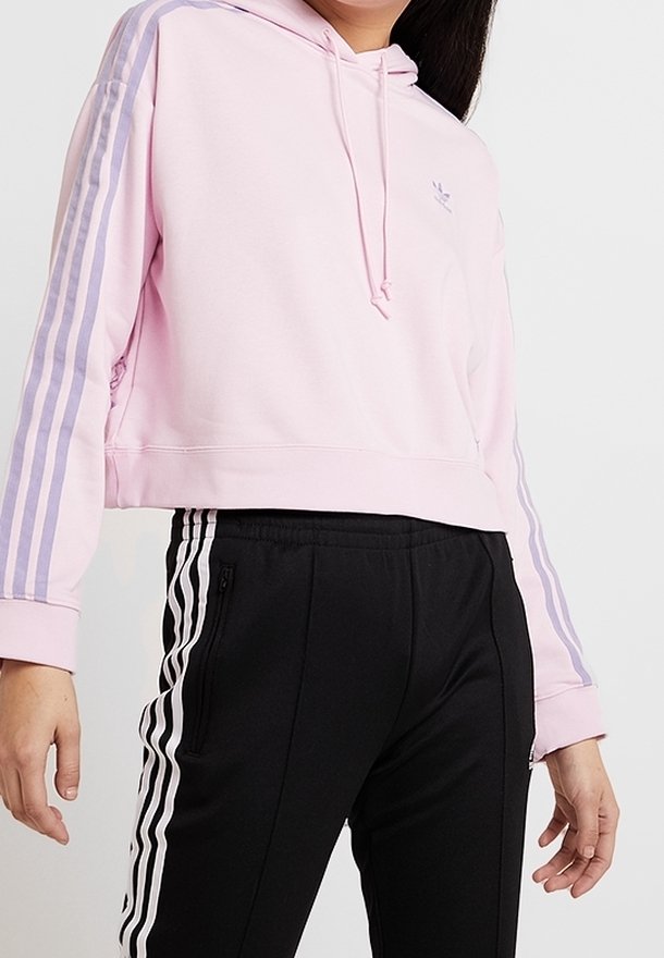 Kvinna iklädd en rosa kortärmad huvtröja med lila ränder och svarta byxor som har vita sidostripes och zip-fickor.