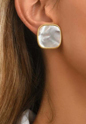 Clou d'oreille carré à bords dorés et aspect perle, porté sur le lobe de l'oreille avec de longs cheveux bruns et une peau lisse visible.