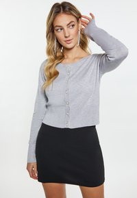 Grå cropped cardigan med lange ærmer, med knaplukning og sølvfarvede knapdetaljer. Sammen med en tætsiddende sort nederdel.