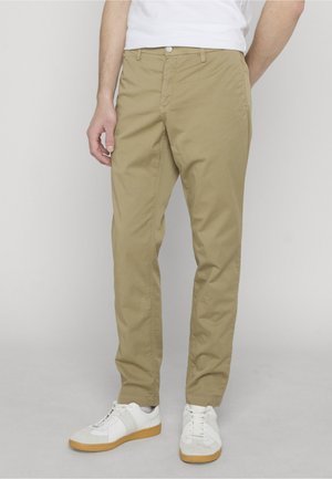 Homme portant un pantalon chino beige et des baskets blanches, debout devant un fond gris clair.
