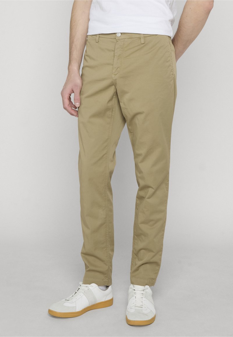 Homme portant un pantalon chino beige et des baskets blanches, debout devant un fond gris clair.
