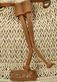 Los cordones de cuero marrón con extremos anudados aseguran una bolsa de paja tejida. Un ojal de metal y una etiqueta "LUNA" destacan el material natural y texturizado.