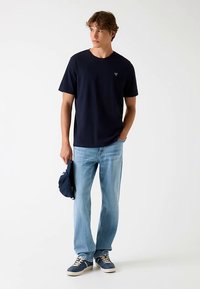 Granatowa koszulka z okrągłym dekoltem z małym logo, w połączeniu z jasnoniebieskimi dżinsami i granatowymi trampkami, trzymająca granatową czapkę w jednej ręce.