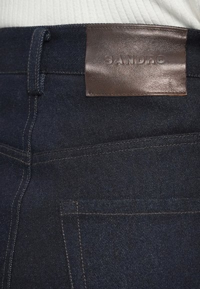 Sötét mosású farmernadrág jellegzetes barna bőr címkével, amelyen az "SANDRO" felirat található. Tartalmazza a standard varrást és zseb részleteket.