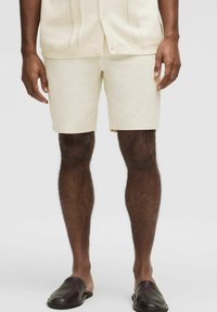 Krémové šortky vyrobené z ľahkej tkaniny, s hladkou textúrou a prispôsobeným strihom, v kombinácii s tmavými koženými šlapkami.