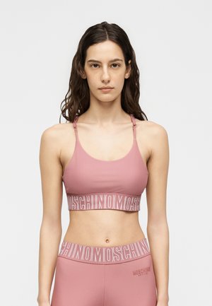 Mladá žena s dlhými tmavými vlasmi oblečená v zladenom ružovom športovom podprsenkovom topu a legínach Moschino na jednoduchom svetlom pozadí.