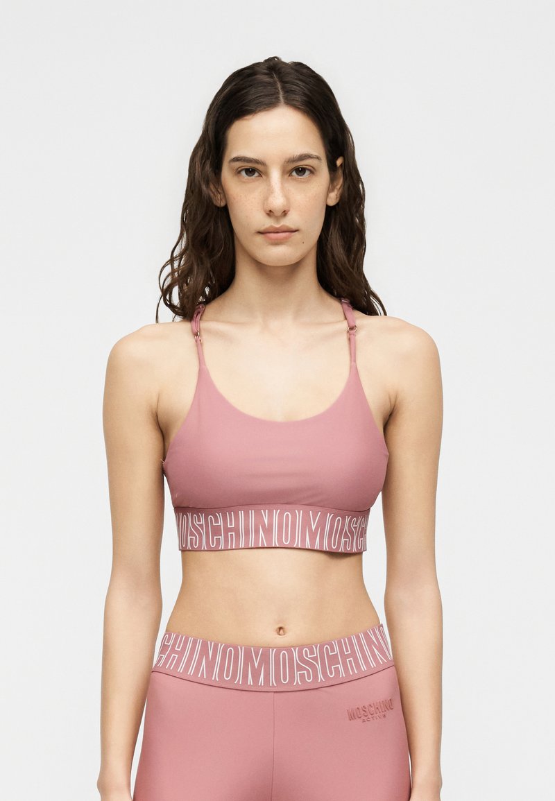 Νεαρή γυναίκα με μακριά σκούρα μαλλιά που φοράει ροζ αθλητικό σουτιέν και κολάν Moschino στον ίδιο τόνο, μπροστά σε απλό φωτεινό φόντο.