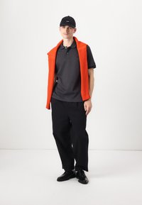 Svart polo skjorte, oransje strikket genser drapert over skuldrene, svarte bukser og svarte sko. Enkelt design med en avslappet passform.