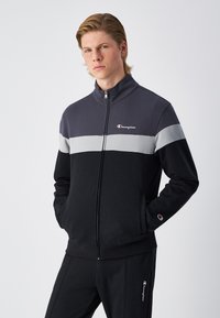 Felpa zip-up nera e grigia con colletto alto, caratterizzata da un design a strisce orizzontali e logo sul petto. Tessuto morbido con tasche.