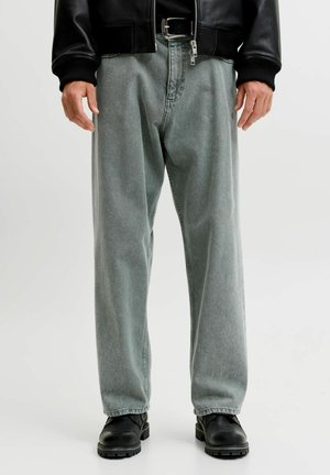 Homme portant un jean gris délavé ample, une ceinture en cuir noire, une veste en cuir noire et des bottes noires à lacets, debout devant un fond uni.