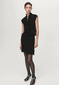 Čierny pletený dvojdielny outfit, ktorý pozostáva z bezrukávového cropped topu s roztrhnutými okrajmi a zodpovedajúcej sukne, doplnený čiernymi silonkami a sandálmi.