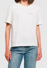 Bílý bavlněný tričko s krátkými rukávy. Má malý růžový grafický motiv srdíčka na prsou a volný střih. Nosené s modrými džíny.