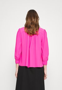 Fuksia blusas su puffed sleeves, gathered back detail, na buttoned cuffs, inayotengenezwa kwa kitambaa laini. Imevaliwa juu ya sketi za mavi.