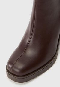Botine din piele maro, cu o textură netedă, vârf rotunjit și o talpă întunecată în contrast. Prezintă un detaliu de cusătură pe lateral.