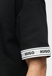 Μαύρο κοντομάνικο με λευκή μανσέτα που φέρει επαναλαμβανόμενο μαύρο κείμενο "HUGO" στο μπράτσο ενός ατόμου σε φωτεινό φόντο.